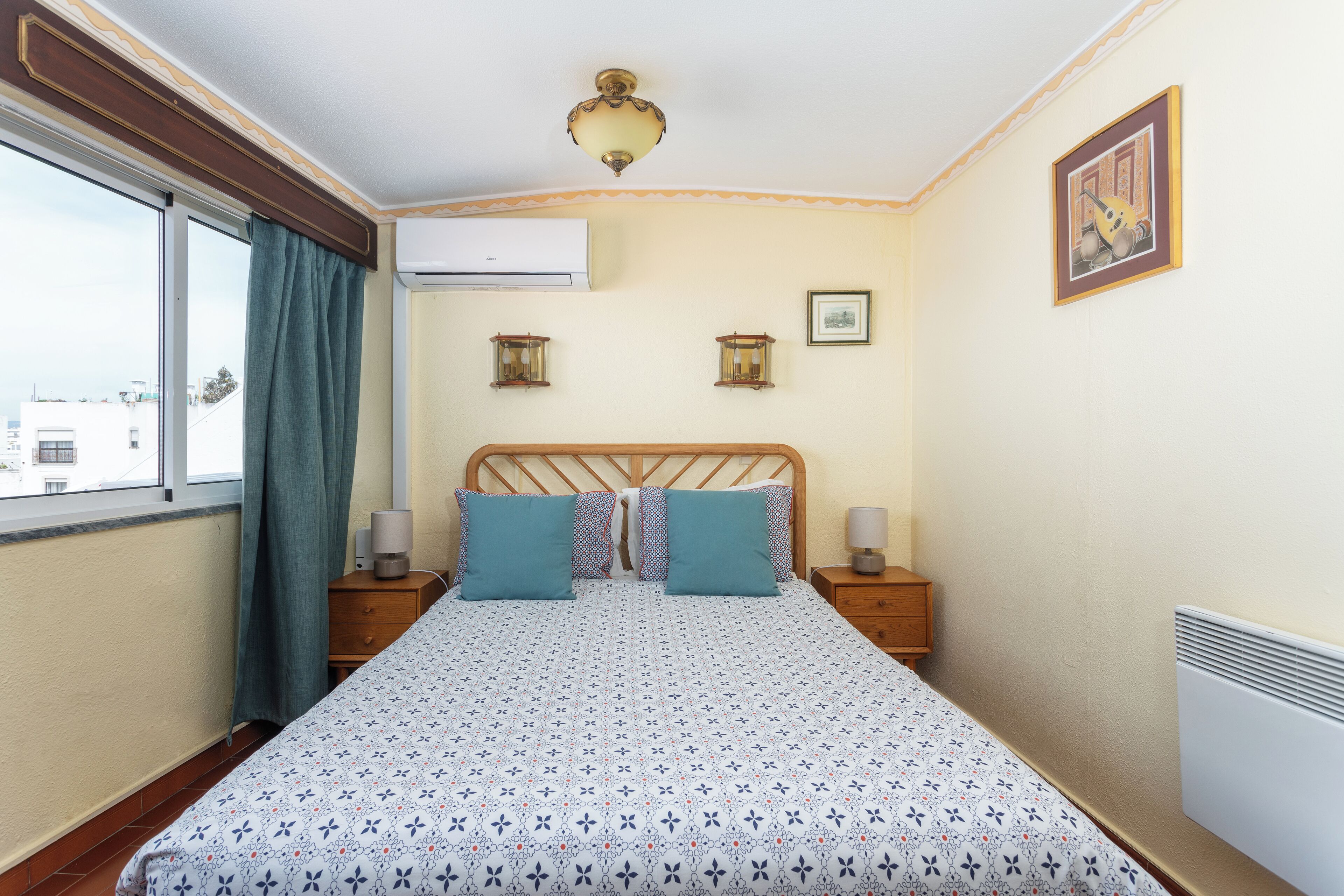 A bedroom with a bed, two bedside tables, a window with curtains, an air conditioning unit, and a wall-mounted light fixture.