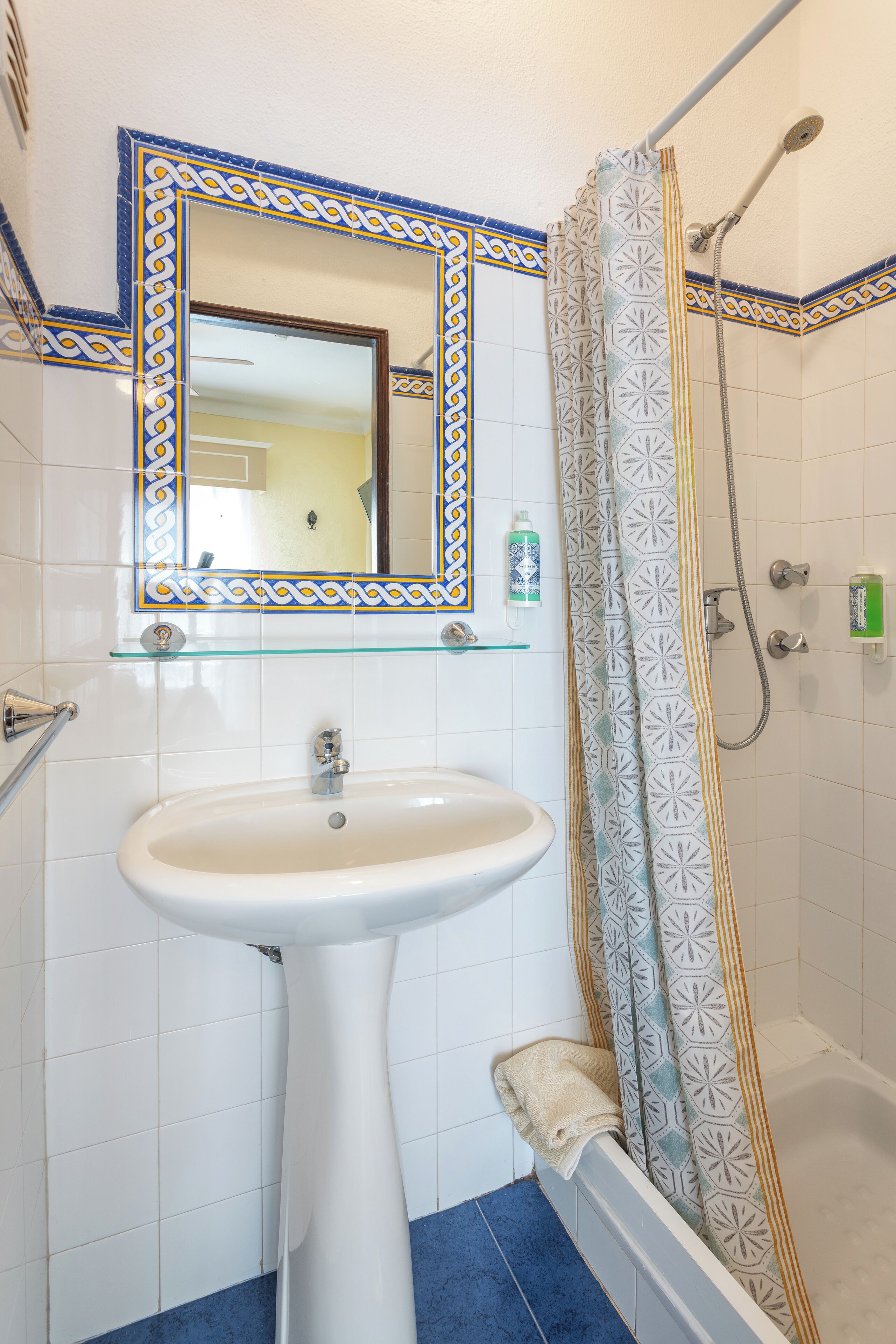 A bathroom with a white sink, a mirror with a decorative frame, a shower with a patterned curtain, and a glass shelf with toiletries.