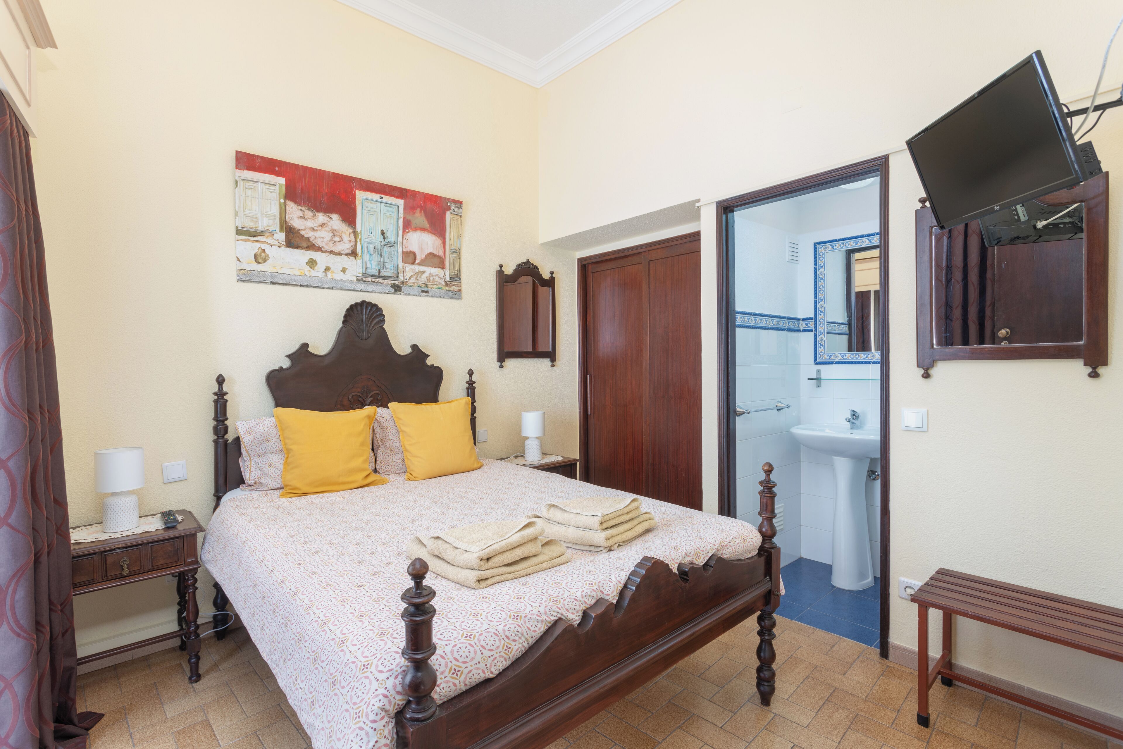 A bedroom with a wooden four-poster bed, a bedside table with a lamp, a painting on the wall, and a television mounted on the wall.