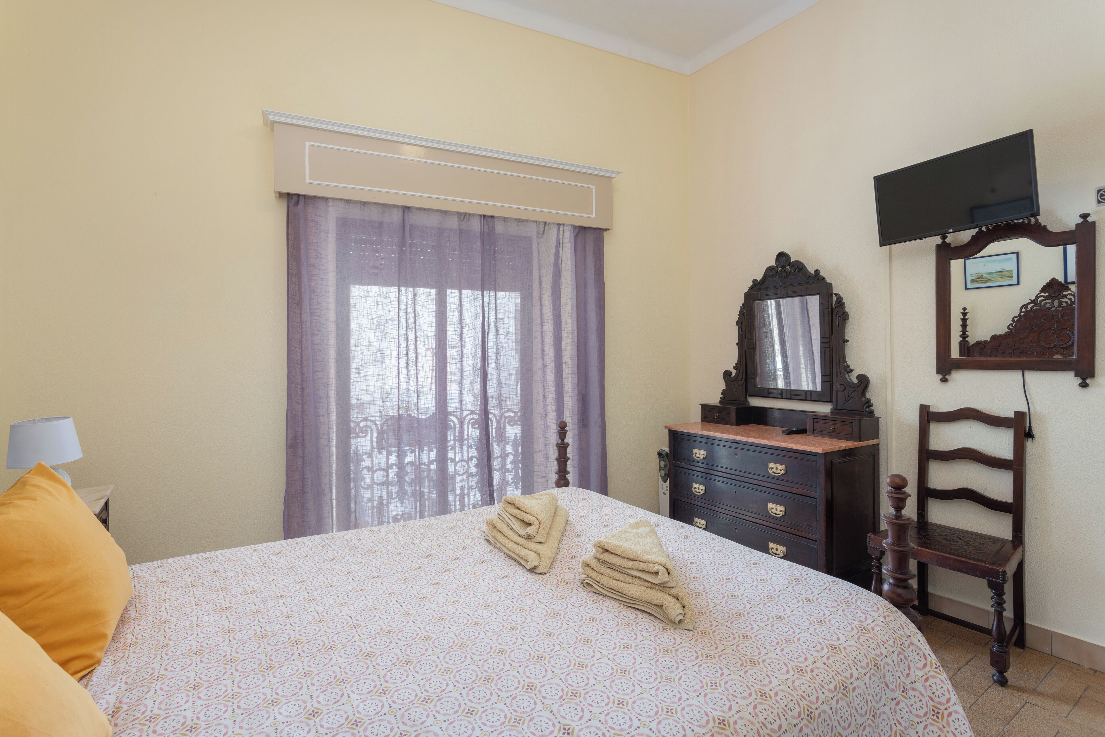 Une chambre à coucher avec un lit, une commode, une chaise, un téléviseur et une fenêtre avec des rideaux.