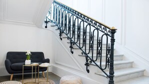 A staircase with ornate black metal railings, a black sofa, two small round ottomans, and a white vase with flowers on a small table. -  Hotel Helzear Etoile (Paris)