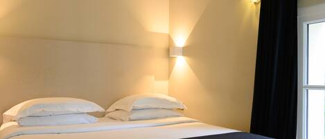 A neatly made bed with white linens and a dark headboard, a wall-mounted light fixture, and a window with black curtains.