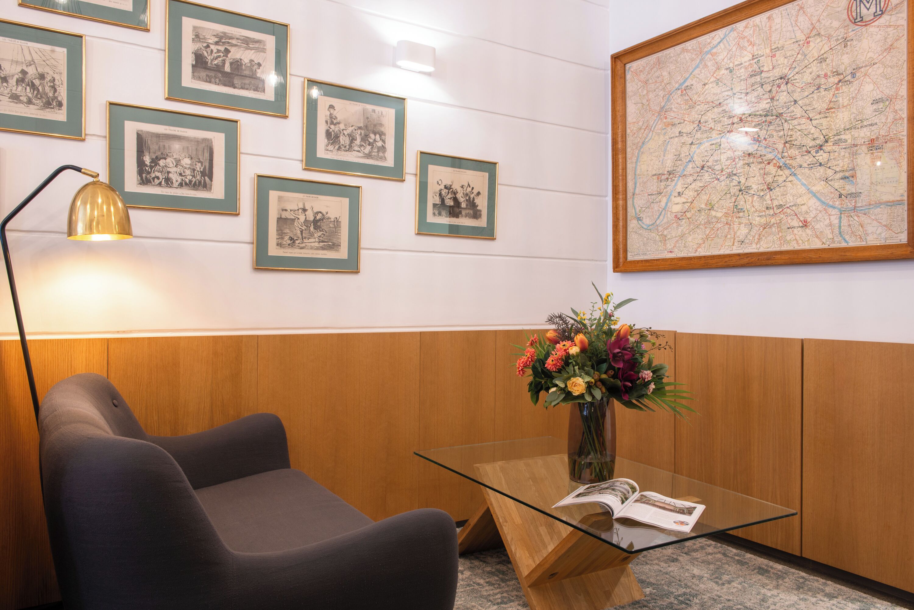 Una habitación con pared de paneles de madera, un mapa, cuadros enmarcados, una mesa de vidrio con un libro, un jarrón con flores y una lámpara.