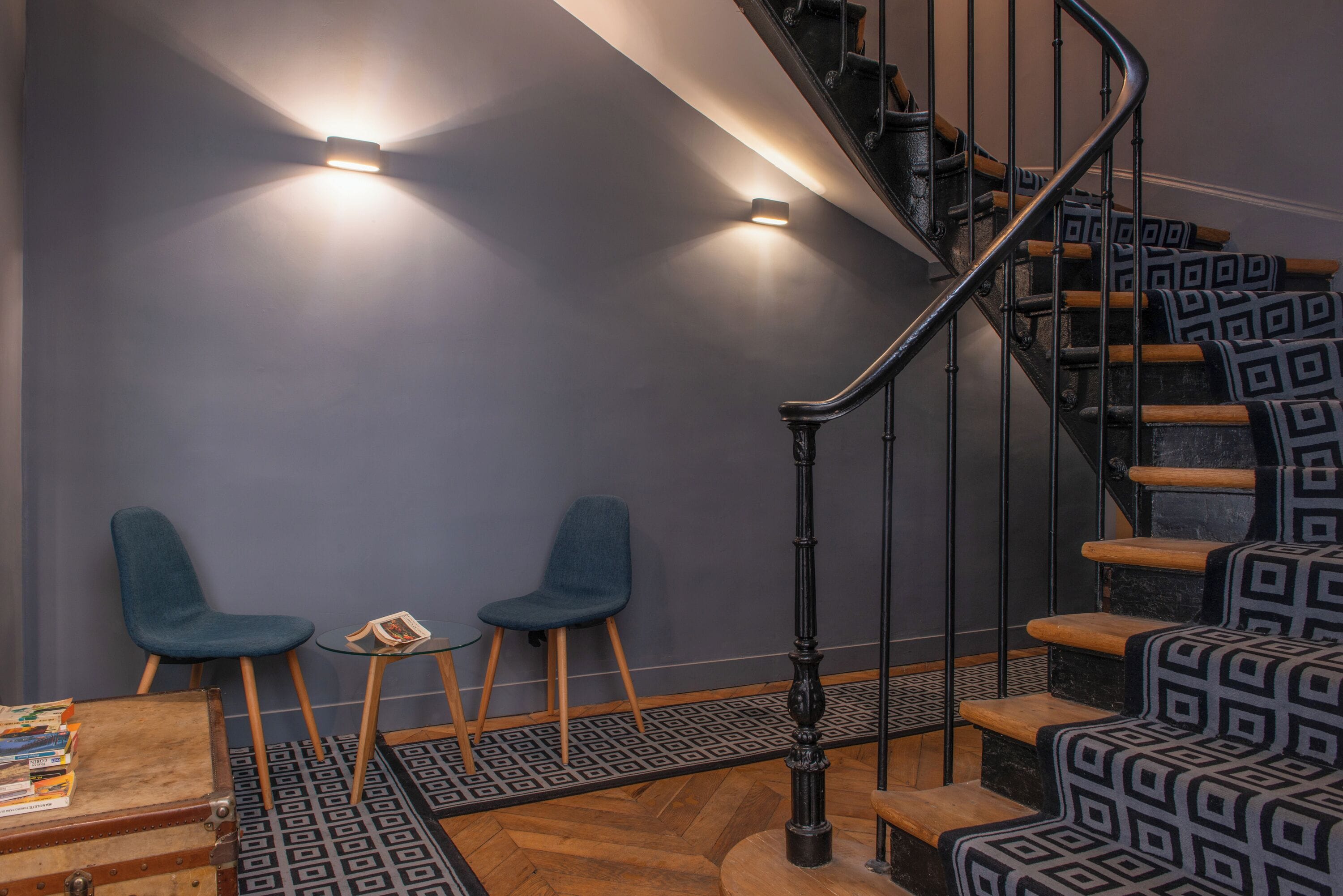 A staircase with patterned steps, two blue chairs, a small table with a book, and a herringbone-patterned floor.
