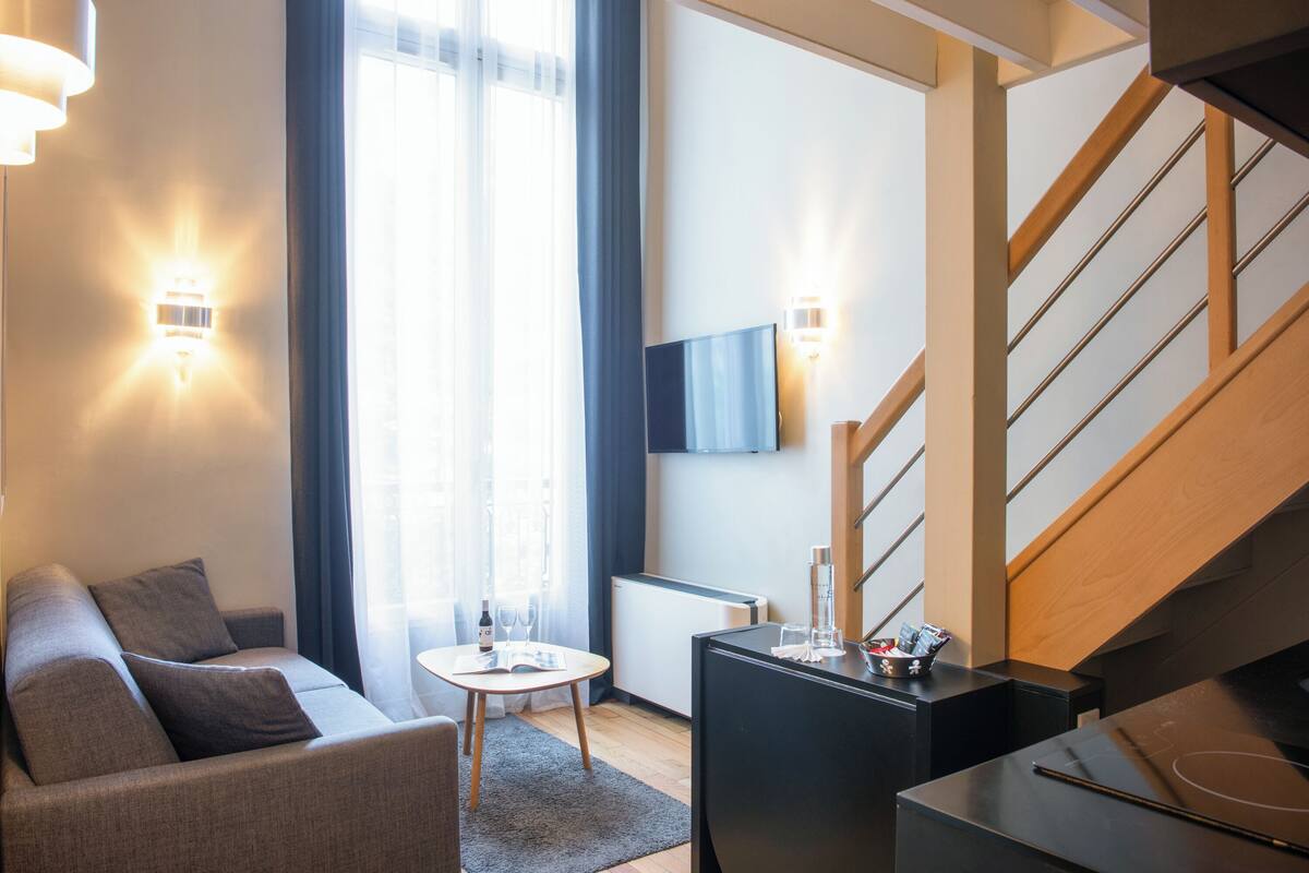 A modern hotel room with a sofa, a small table, a TV mounted on the wall, and a staircase.