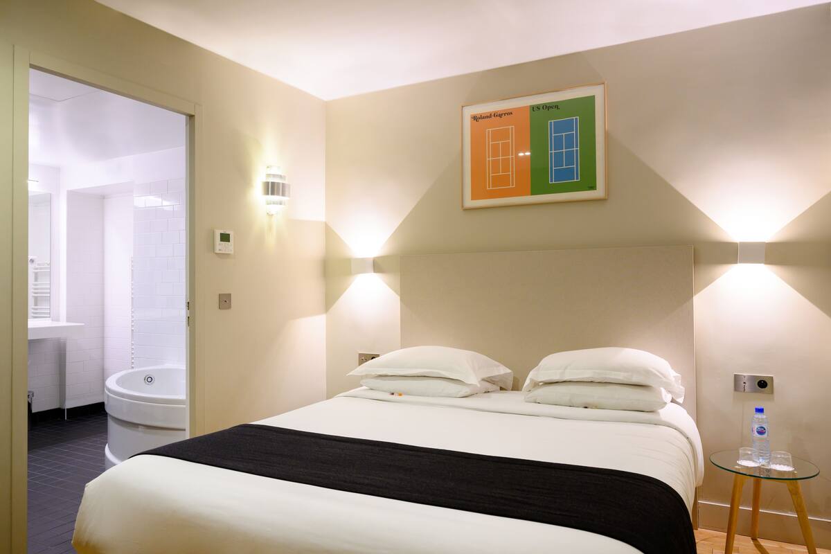 A hotel room with a bed, a glass side table, a wall-mounted light, a framed picture, and a bathroom visible through an open door.