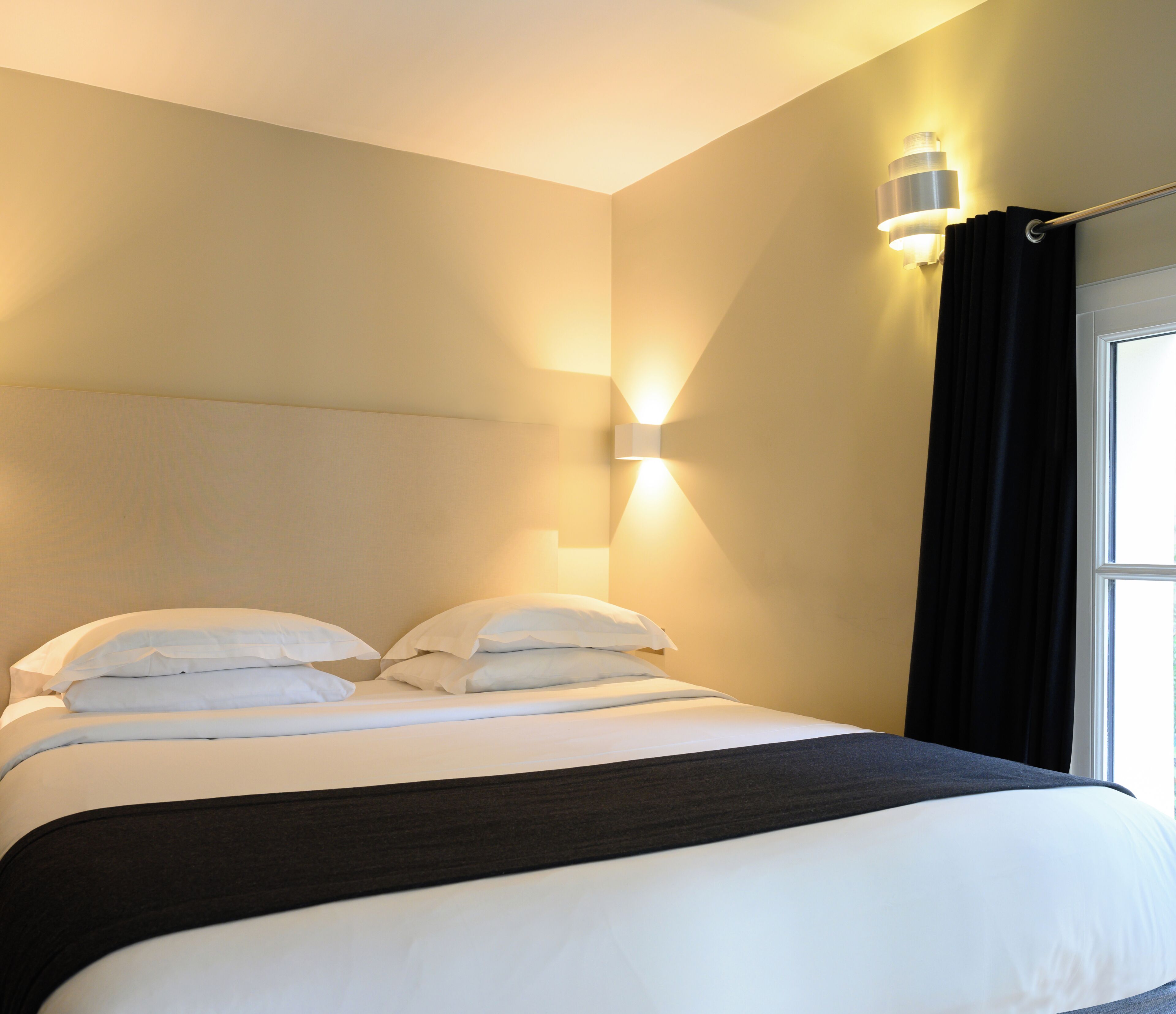 A neatly made bed with white linens and a dark headboard, a wall-mounted light fixture, and a window with black curtains.