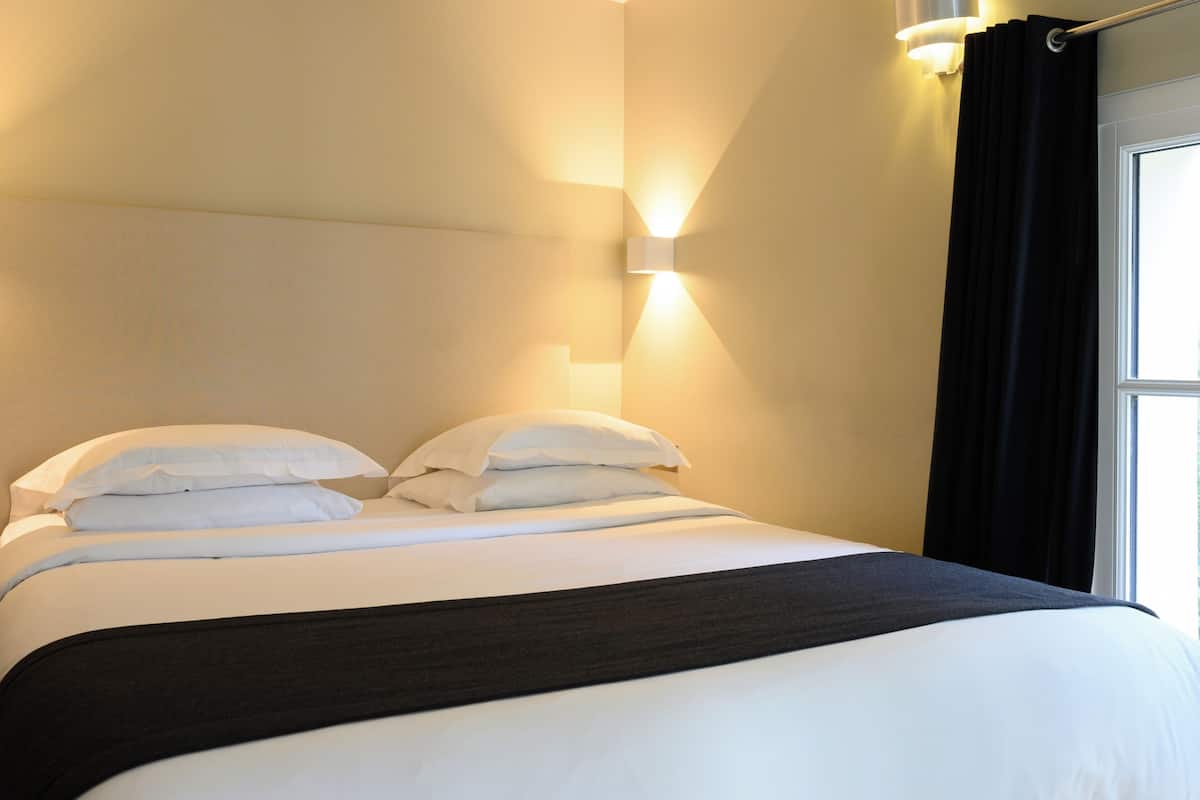 A neatly made bed with white linens and a dark headboard, a wall-mounted light fixture, and a window with black curtains.