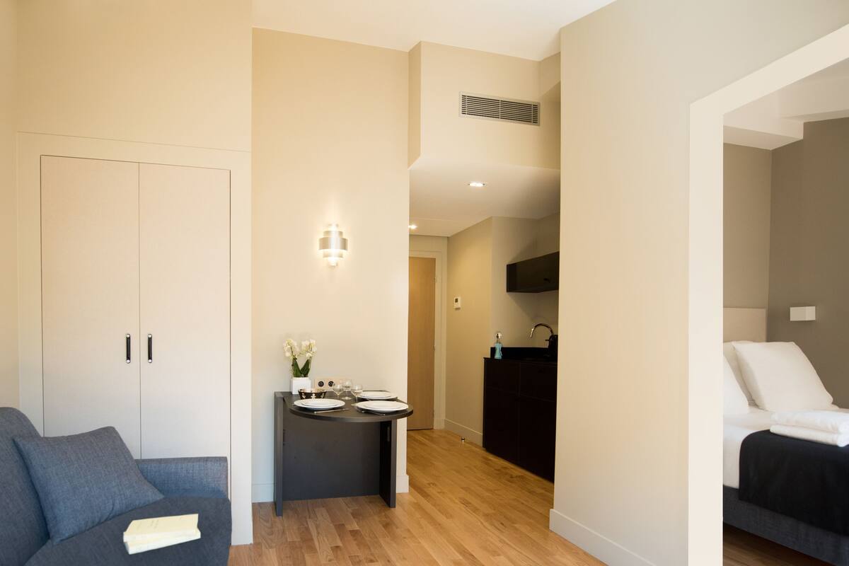 A modern apartment with a grey sofa, a dining area with a small table, a kitchen, and a bedroom with a bed.