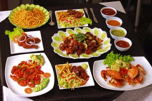 Uma mesa com diversos pratos, incluindo macarrão, frango frito, camarão e caranguejo, acompanhados de molhos e guarnições variadas.
