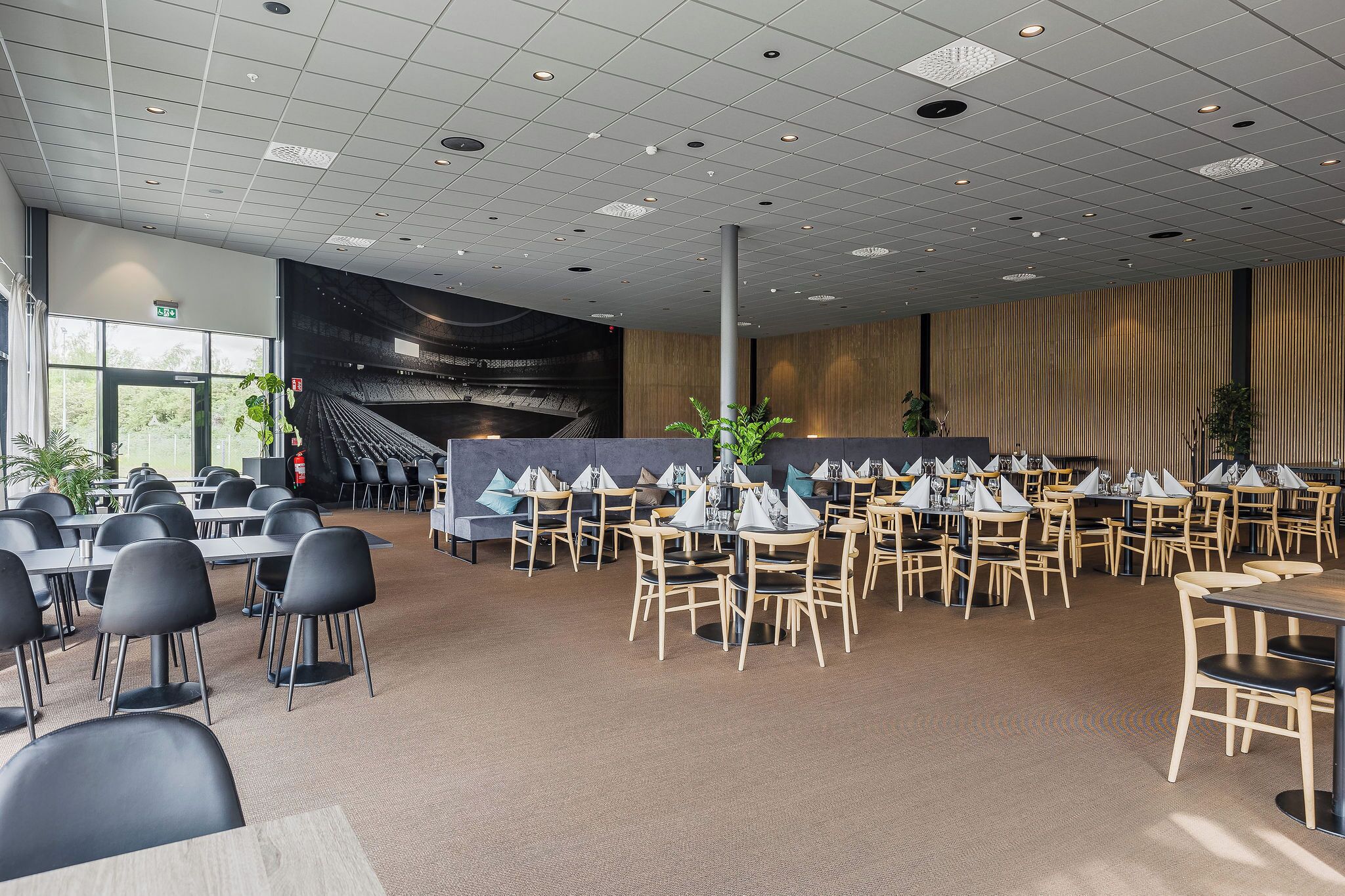 A spacious indoor dining area with multiple tables and chairs, a high ceiling with recessed lighting, and a large window providing natural light.