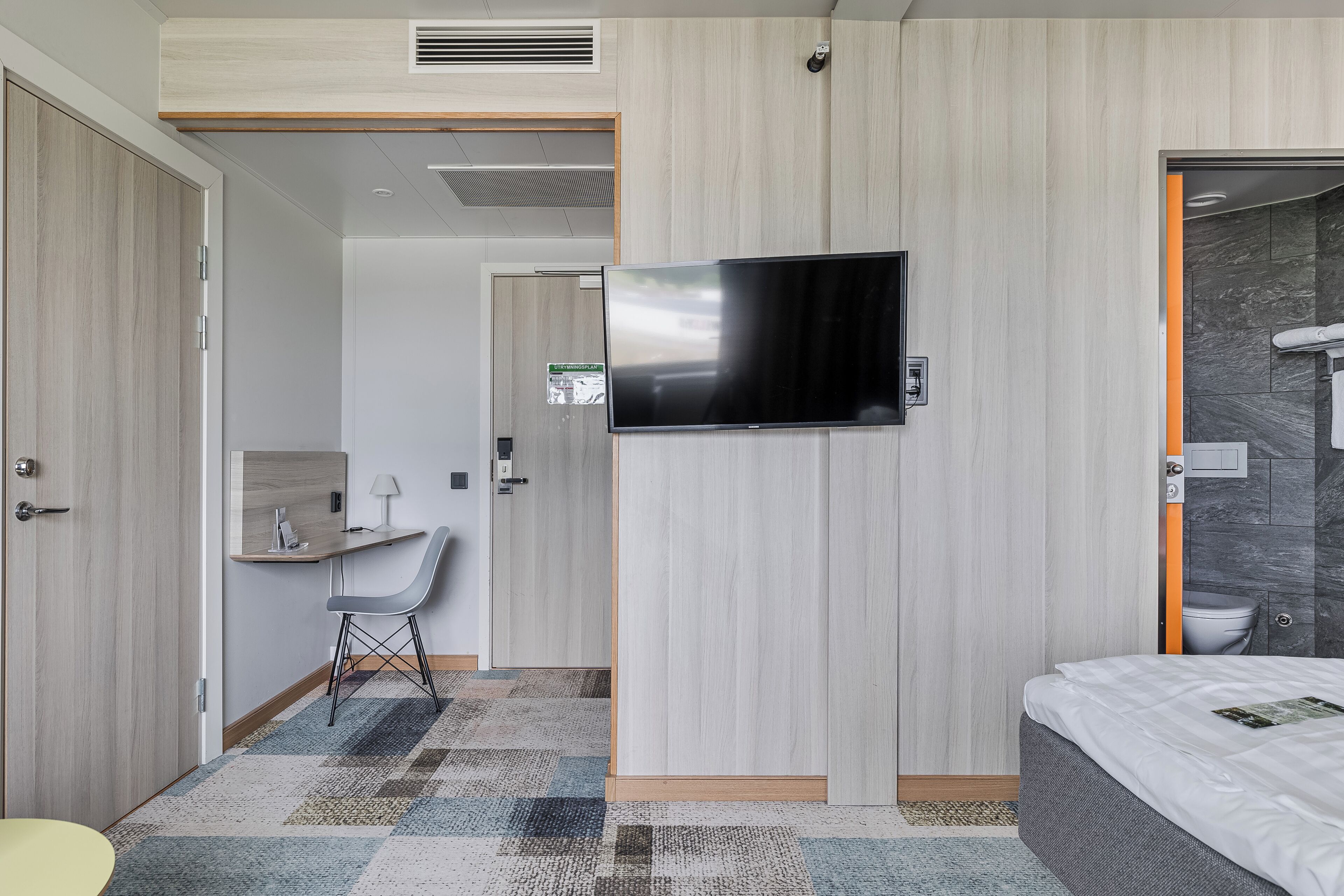 A modern hotel room with a flat-screen TV, a desk with a chair, a bathroom with a shower, and a bed.