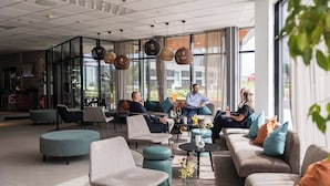 A modern lounge area with sofas, chairs, and tables, large windows with curtains, and hanging spherical light fixtures. - Arenahotellet i Uppsala (Uppsala)
