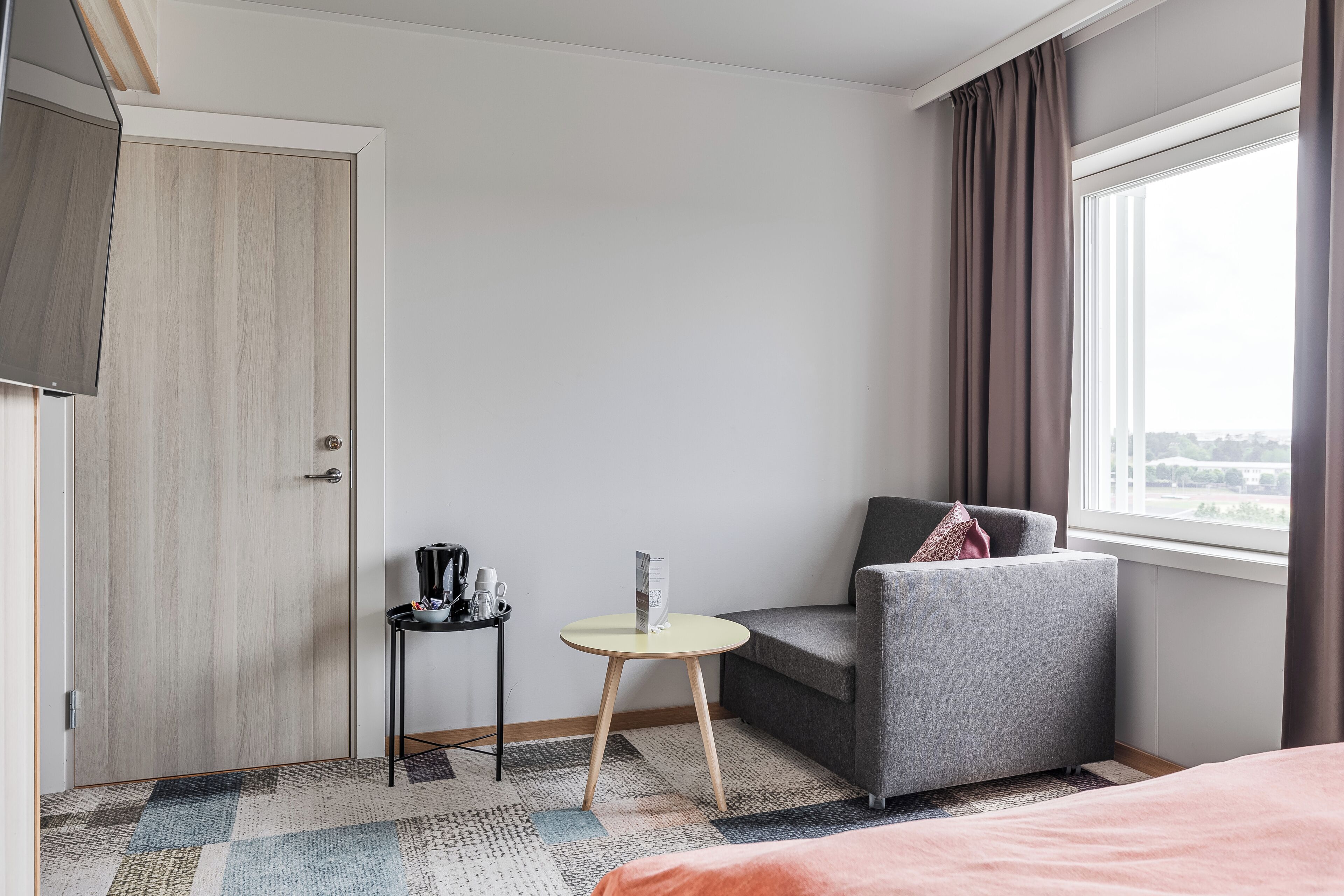 A hotel room with a bed, a grey armchair, a small table with a kettle and a magazine, a door, and a window with curtains.