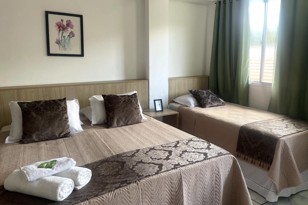 A hotel room with two beds, each with a brown bedspread and white pillows. There is a framed picture on the wall, a nightstand with a lamp, and a window with green curtains.