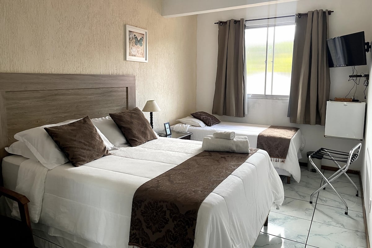 A hotel room with two beds, a flat-screen TV, a refrigerator, and a window with curtains.
