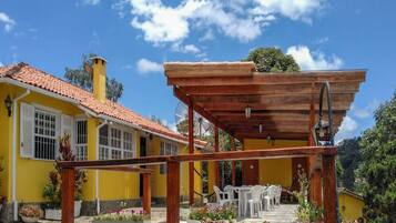 Un edificio amarillo con techo de tejas rojas, una pérgola de madera y mesas al aire libre.