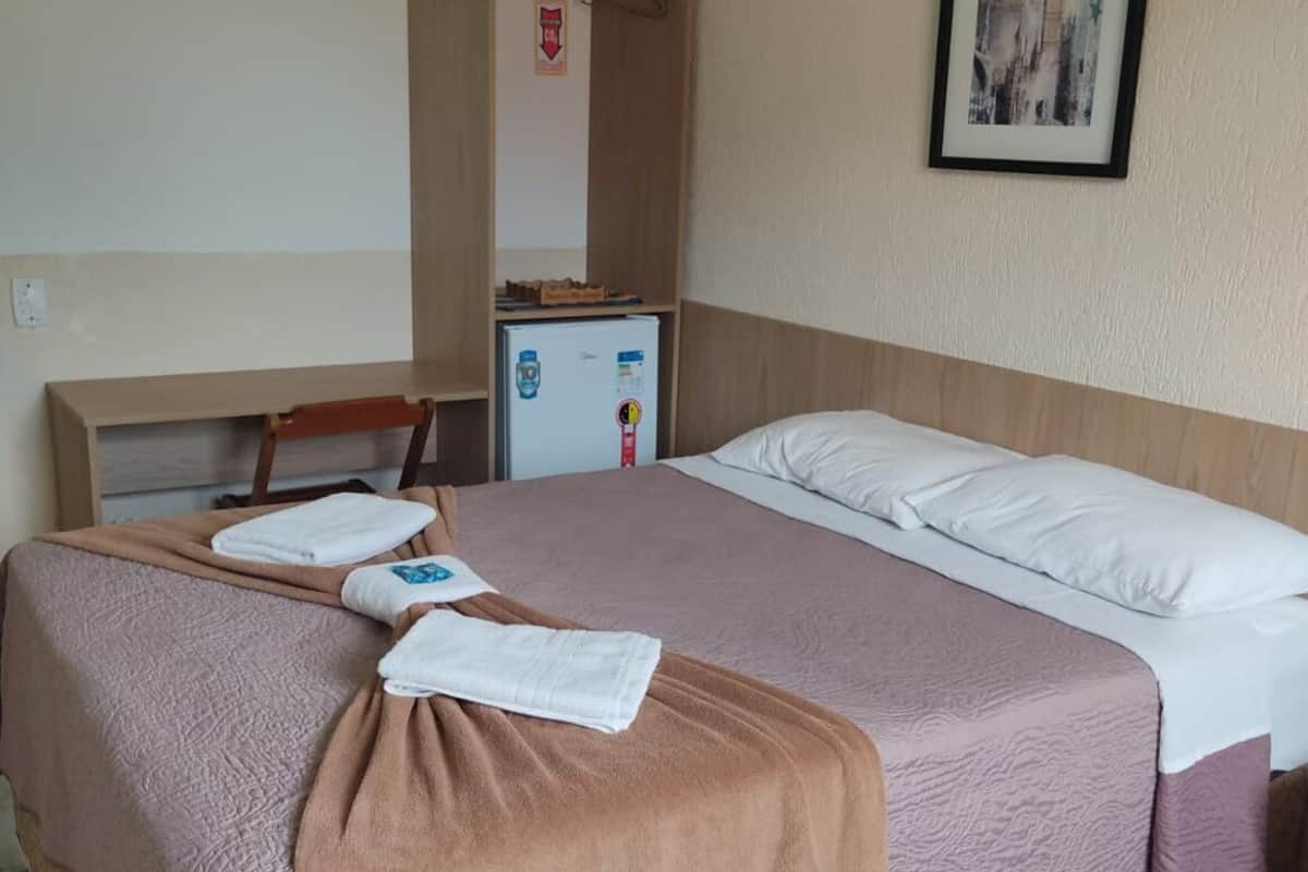 A hotel room with a bed, a desk, a chair, a wardrobe, a small fridge, and a framed picture on the wall.