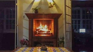 A fireplace with a fire, a table with a patterned tablecloth, and a potted plant on the mantel. - Pousada Sítio Sossego (Petrópolis)