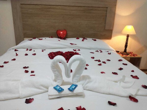 A bed with rose petals and a heart-shaped balloon, a white towel with blue heart-shaped decorations, and a bedside lamp.