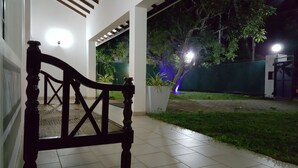 A covered outdoor area with a wooden bench, a tiled floor, and a well-lit garden with plants and a fence. - Tripper's Private Villa (Negombo)
