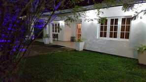 Un edificio blanco con un césped bien cuidado, plantas grandes en macetas y una ventana con cortinas.