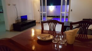 A dining area with a wooden table set for two, a television on a stand, and a view of a pool area through glass doors. - Tripper's Private Villa (Negombo)