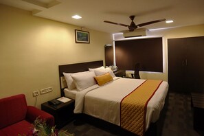 Quarto de hotel com cama, poltrona vermelha, mesa de cabeceira com telefone e ventoinha no teto.