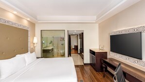 A hotel room with a large bed, a TV, a desk, and a bathroom visible through a glass door.