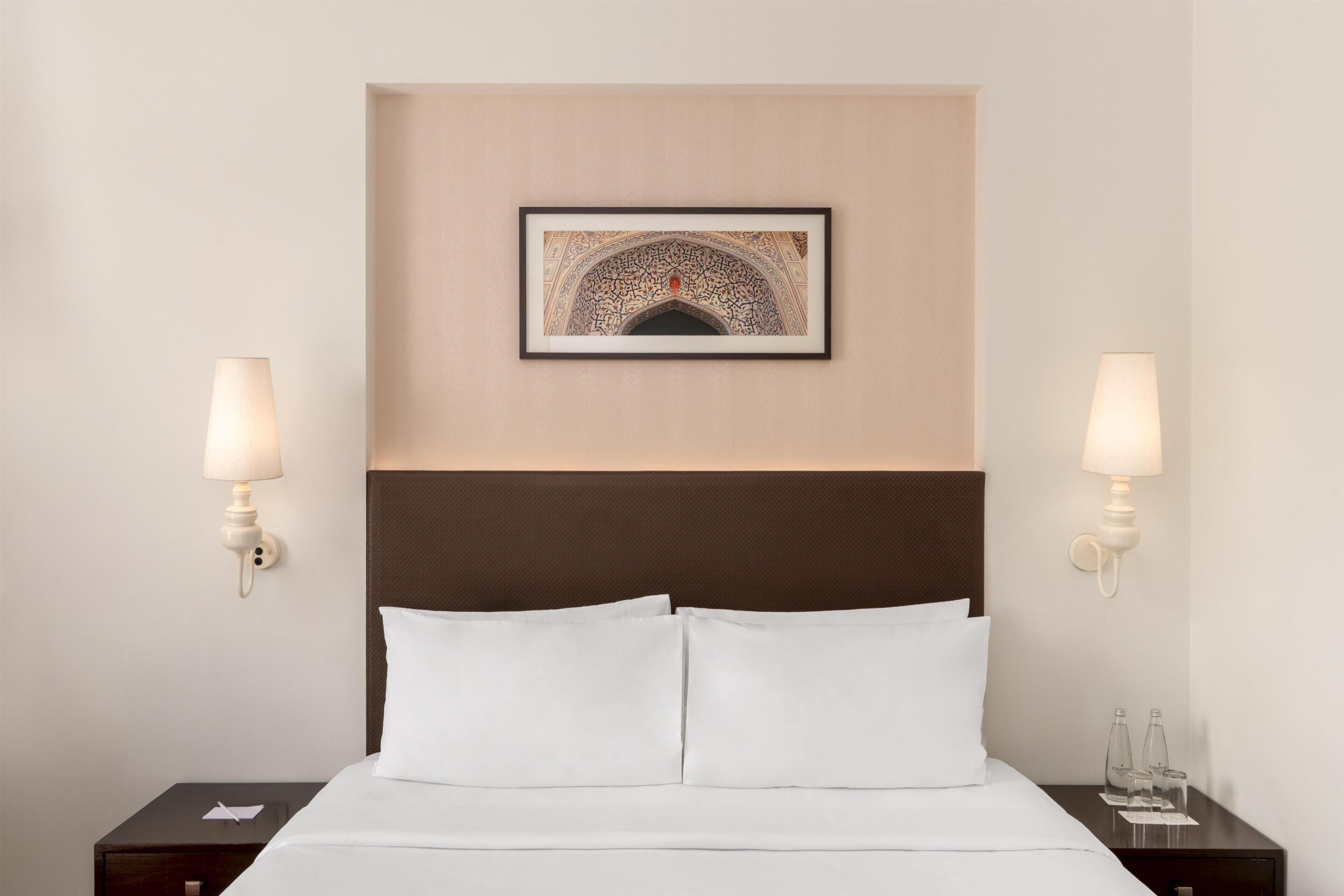 A hotel room with a bed, two bedside lamps, a framed artwork above the bed, and a nightstand with a glass of water.