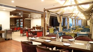 A hotel dining area with multiple tables and chairs, a buffet setup, and a view of the cityscape through large windows.
