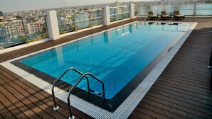 A rooftop infinity pool with a wooden deck, lounge chairs, and a city view.