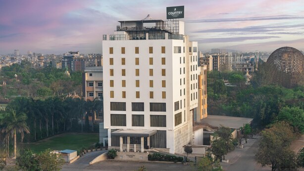 A modern hotel with a prominent sign, surrounded by greenery and other buildings.