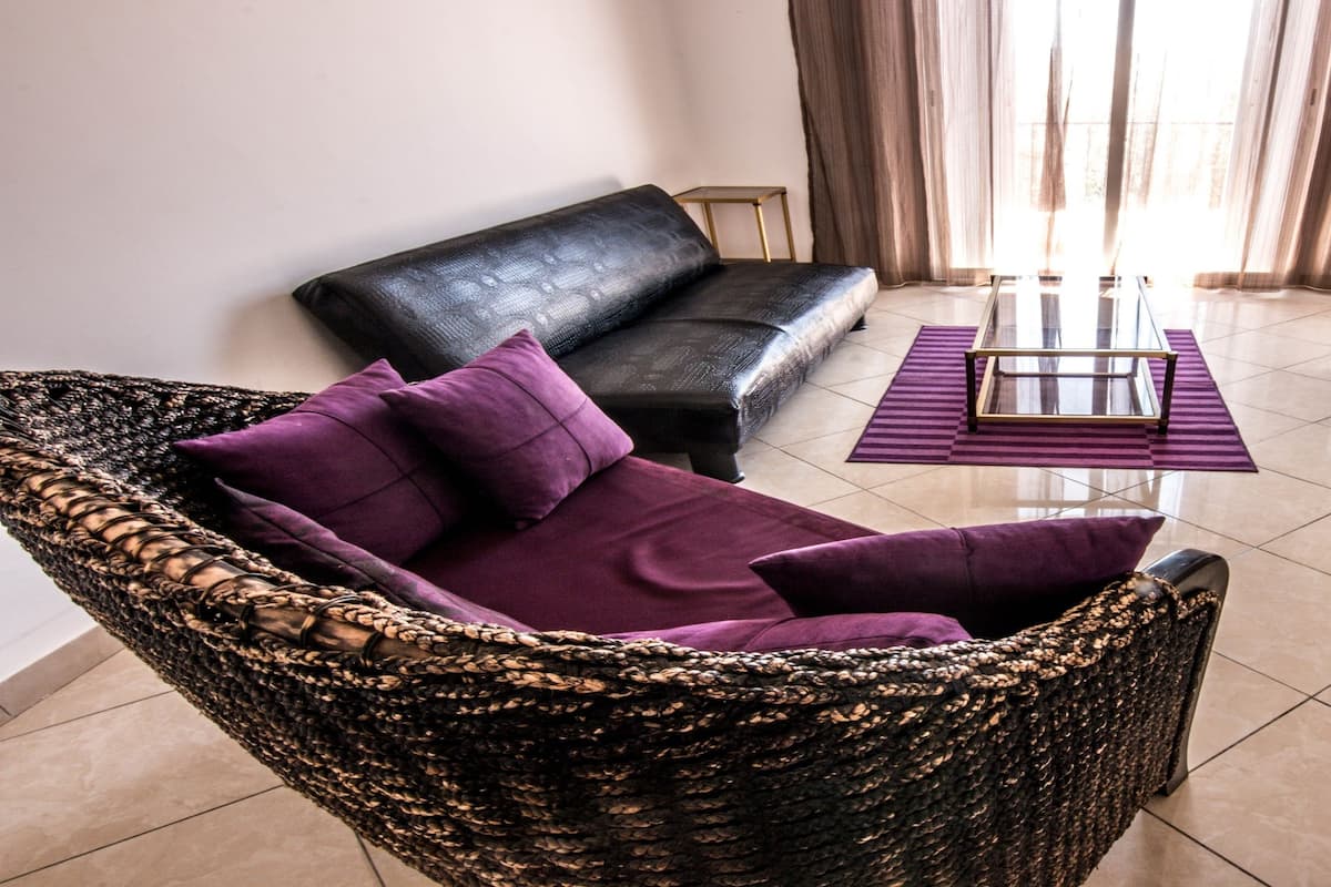 A living room with a black sofa, a wicker chair, a glass coffee table, and a television.