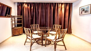 A dining area with a round glass table surrounded by six wicker chairs with white cushions, a wicker sideboard, and a large window with dark curtains. - Mahdia Place Apartments (Mahdia)