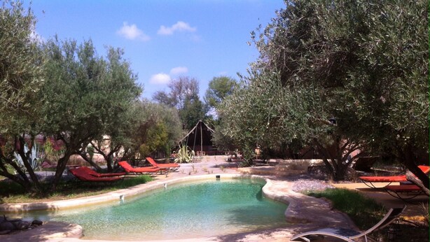 Zona de piscina con sillones rojas, rodeada de árboles y un pequeño puente.