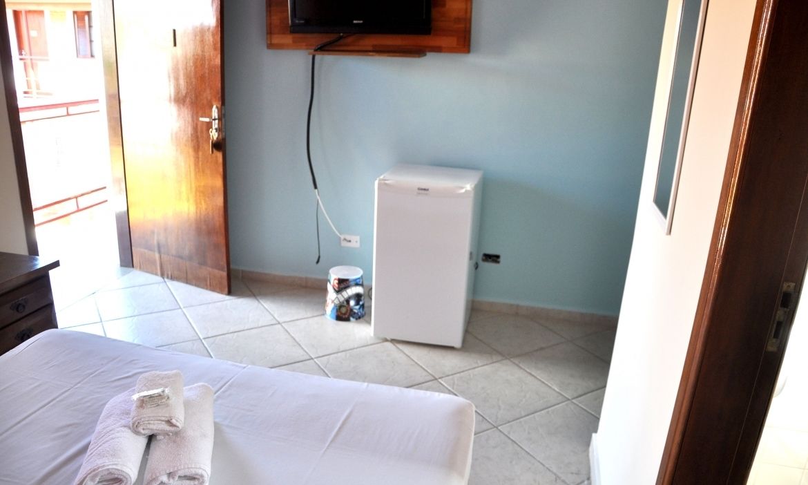 Quarto de hotel com uma cama, um pequeno frigorífico branco, uma televisão fixada na parede e chão em azulejo.
