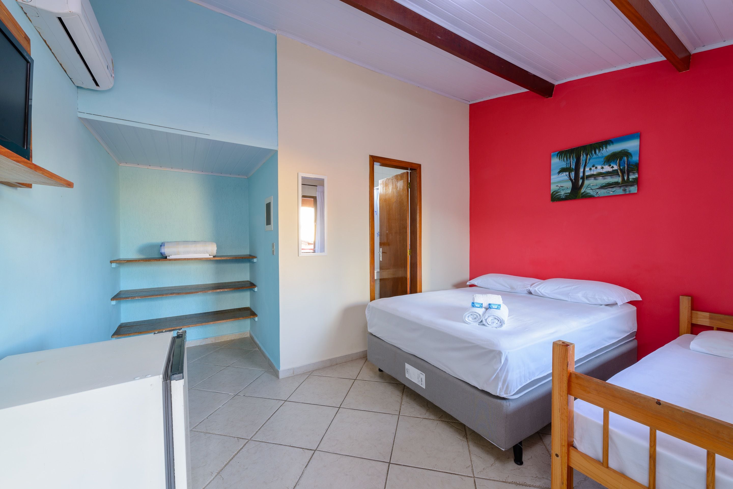 A room with a bed, a wooden chair, a shelf with towels, a painting on the wall, and a door.