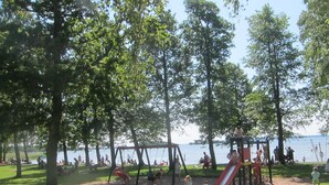 A park with playground equipment, including swings and a slide, surrounded by trees and a body of water in the background. - Sikhalls Camping och Restaurang (Bralanda)