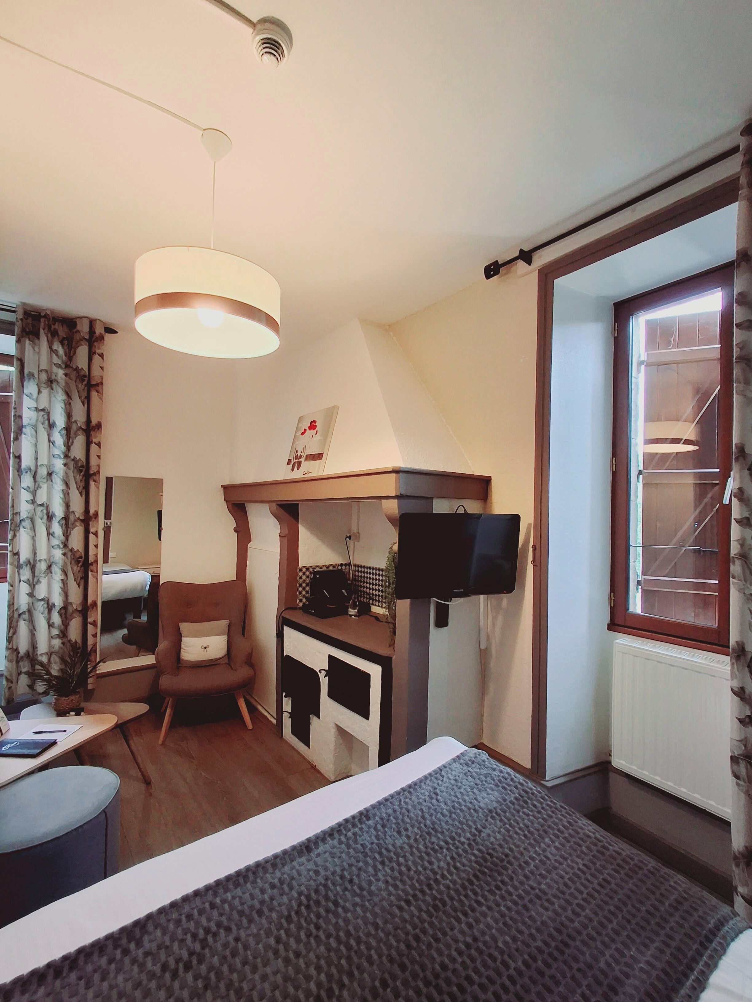 A hotel room with a bed, a desk with a computer, a chair, a window with a view of a building, and a ceiling light.