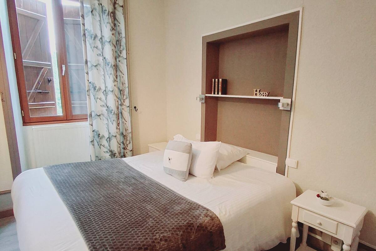 A neatly made bed with a brown blanket, a bedside table with a bowl, and a shelf with books and a sign.