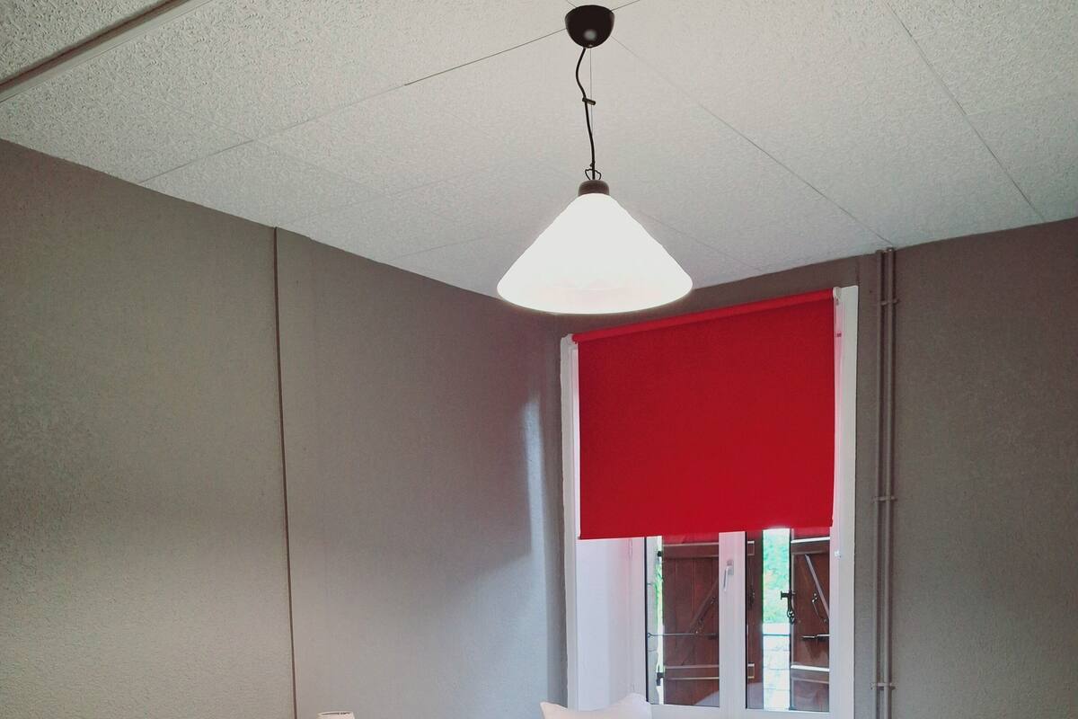A room with two beds, a window with a red blind, a ceiling light, and a bedside table with a lamp.