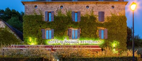 Hotel La Brasserie features a classic stone building with a flat roof and ivy-adorned facade, set amidst a landscaped area with a stone wall and potted plants.