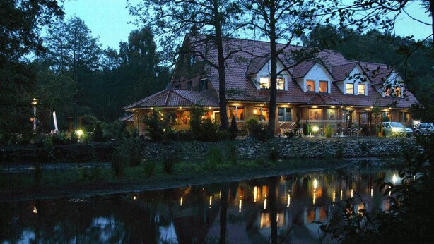 Une grande maison à deux étages, dotée d’un toit en tuiles, entourée d’arbres et bordée par une étendue d’eau dans laquelle se reflète le bâtiment.