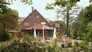 Un bâtiment en briques surmonté d’un toit en tuiles, avec un espace extérieur aménagé avec des tables et des parasols, le tout entouré de verdure et de rochers.