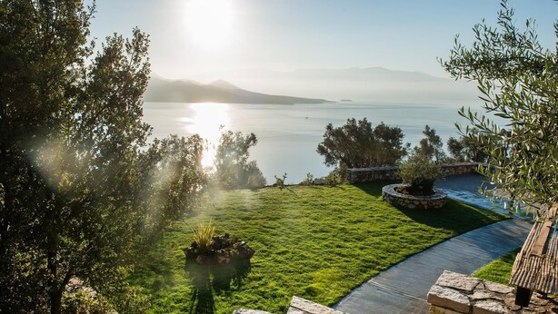 A landscaped garden with a stone pathway leading to a body of water, surrounded by trees and a view of distant mountains. - Pearl Mansions (Lefkada)