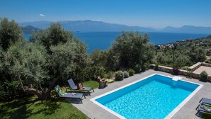 A rectangular swimming pool with a clear blue pool, surrounded by a stone deck and a grassy area with trees. - Pearl Mansions (Lefkada)