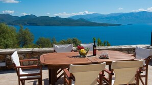 An outdoor dining area with a table and chairs, a bowl of fruit, and a bottle of wine. - Pearl Mansions (Lefkada)