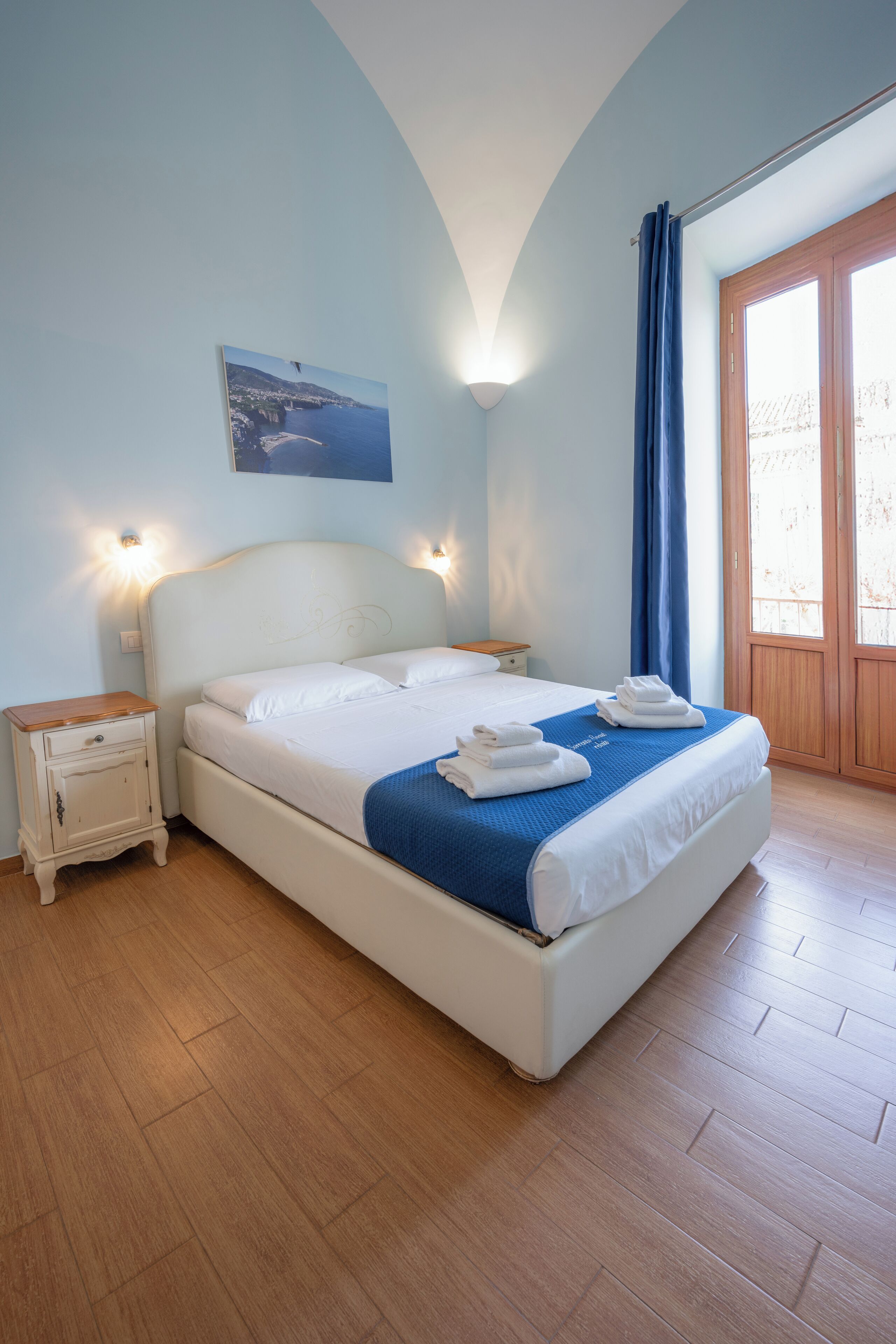 A neatly made bed with a blue blanket and towels, a nightstand with a lamp, and a view of the sea through a window with curtains.