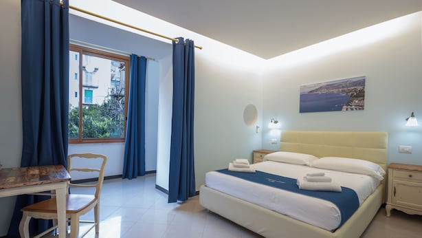 A bedroom with a bed, a desk, a chair, a window with curtains, a bedside table, a nightstand, and a wall-mounted lamp. - Sorrento Coast Relais (Meta)